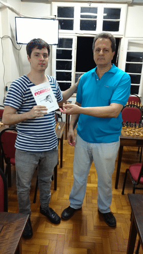 Leo Ramos Simões recebendo das mãos de Fernando Madeu o livro prêmio que ganhou pela conquista do Rápido FIDE de Agosto. Tal livro foi uma doação do poeta Ricardo França, recém associado à ALEX.