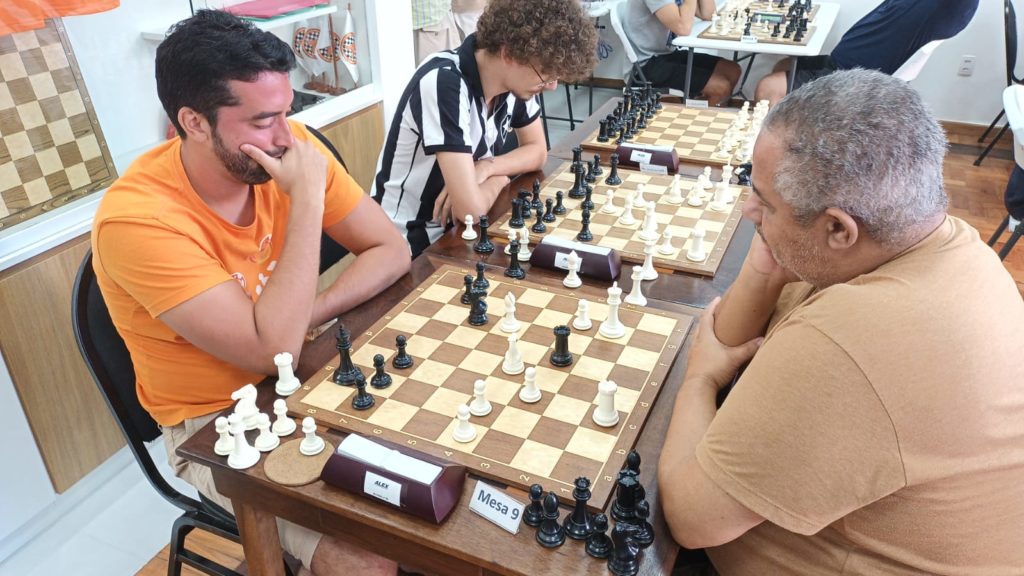 Interior do clube de xadrez com tabuleiro, peças e relógio.